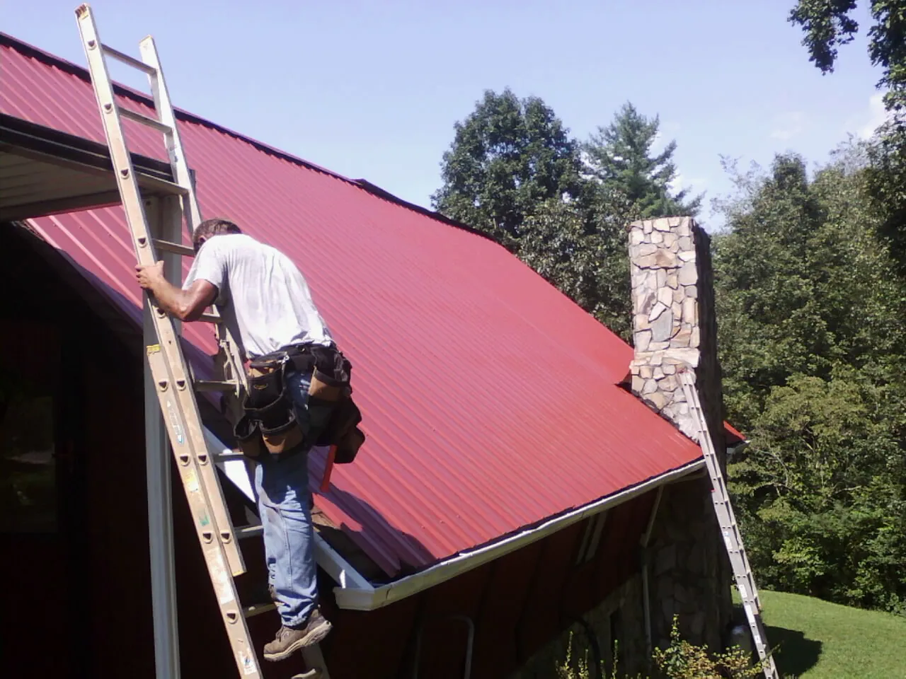 Roofer on a ladder beside a red metal roof and stone chimney — Soffit & Fascia Repair by Roofing Brothers in Eugene
